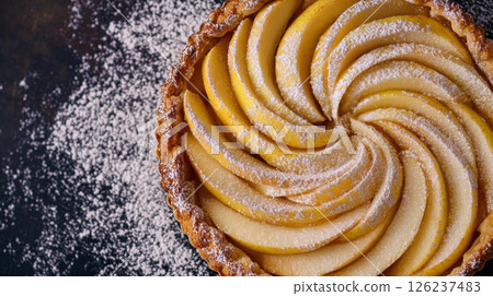 A beautifully arranged apple tart features thin slices of apple in a spiral design, lightly dusted with powdered sugar on a dark background. 126237483