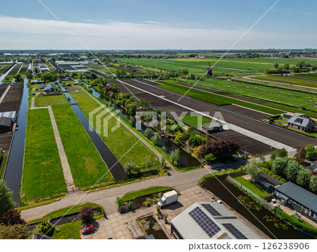 Aerial view of a lush countryside garden surrounded by canals and farmlands, with a cozy house and colorful trees, set in a peaceful rural landscape under a clear blue sky. 126238906