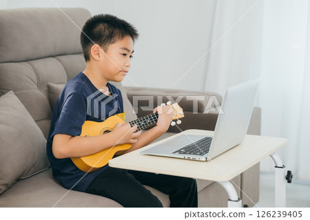 Boy learning ukulele on a sofa while watching a tutorial on a laptop. The cozy living room scene highlights the integration of technology and creative education in a modern home environment. 126239405