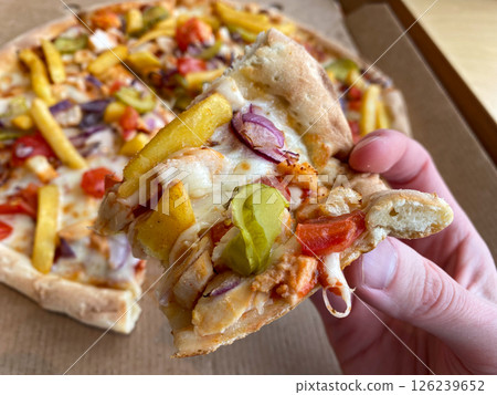 A man holds a slice of pizza in his hand, a hearty pizza with vegetables, chicken and fries 126239652