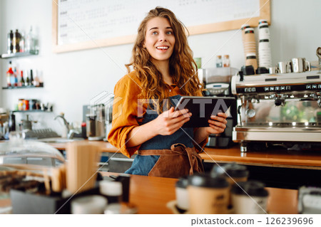 Smiling female barista takes an order from a tablet while standing at bar counter in a coffee shop. Smiling female barista takes an order from a tablet while standing at bar counter in a coffee shop. 126239696