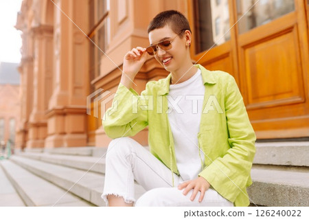 Positive woman with a short haircut and a green shirt walks and relaxes on the streets of the city. Positive woman with a short haircut and a green shirt walks and relaxes on the streets of the city. 126240022