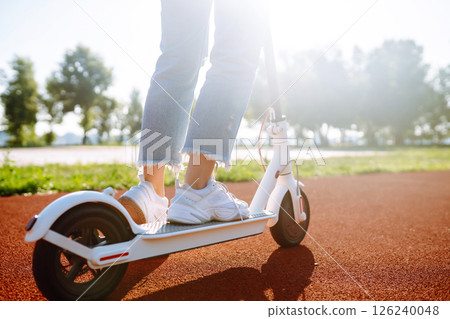 Woman riding electric kick scooter outdoors at sunset, closeup. Ecological transportation concept. Woman riding electric kick scooter outdoors at sunset, closeup. Ecological transportation concept. 126240048