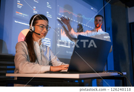 Stock broker is sitting and working by laptop. Man is standing near her, showing data on projector Stock broker is sitting and working by laptop. Man is standing near her, showing data on projector 126240118