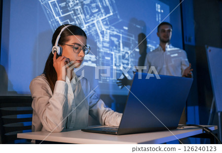 Stock broker is sitting and working by laptop. Man is standing near her, showing data on projector 126240123