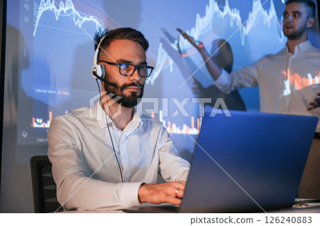 Stock broker is sitting and working by laptop. Man is standing near him Stock broker is sitting and working by laptop. Man is standing near him 126240883