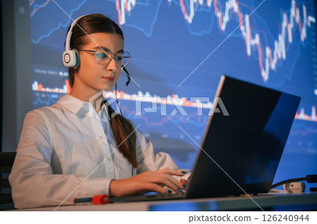 Sitting by laptop. Female stock broker is working indoors. Big wide display behind her 126240944