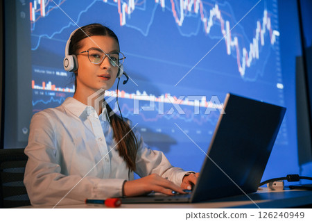 In headphones, with laptop. Female stock broker is working indoors. Big wide display behind her 126240949