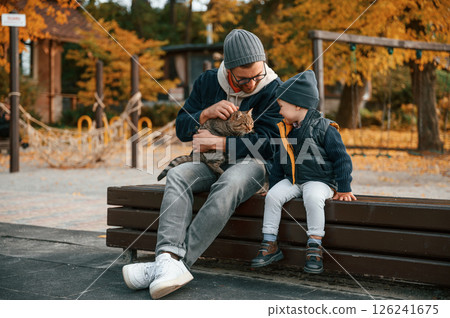 Sitting with cute cat. Father and young son is together outdoors at daytime Sitting with cute cat. Father and young son is together outdoors at daytime 126241675