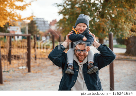 Holding boy on the shoulders. Father and young son is together outdoors at daytime Holding boy on the shoulders. Father and young son is together outdoors at daytime 126241684