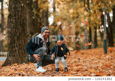 Playing with fallen leaves. Father and young son is together outdoors at daytime Playing with fallen leaves. Father and young son is together outdoors at daytime 126241862