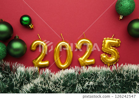 Numbers of year 2025 made by gold candles with christmas green toy on red background. Top view 126242547