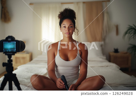 A relaxed young woman sits on a comfortable bed holding a camera device, set in a cozy and well-decorated bedroom with soft lighting. Ideal for influencer and lifestyle themes. 126249028
