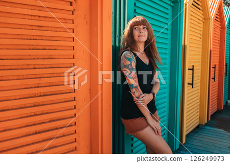 Young smiling woman with a tattoo on her arm, standing leaning by beach's changing room. Multicolored wooden houses on the background. The concept of summer vacation 126249973