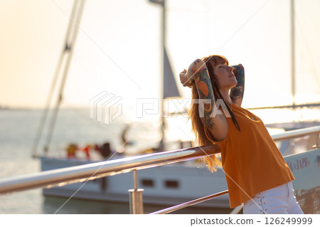 Pretty tanned young woman with tattoos relaxed leaning on back on railing of ship, closed eyes. Copy space. Sea cruise and summer vacation. Concept of freedom Pretty tanned young woman with tattoos relaxed leaning on back on railing of ship, closed eyes. Copy space. Sea cruise and summer vacation. Concept of freedom 126249999
