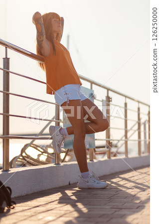 Smiling tanned young woman with tattoos relaxed leaning on back on railing of ship. Vertical orientation. Sea cruise and summer vacation. Concept of freedom Smiling tanned young woman with tattoos relaxed leaning on back on railing of ship. Vertical orientation. Sea cruise and summer vacation. Concept of freedom 126250000