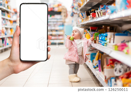 Shopping. A small child stands at the supermarket shelves with a pear in his hand. The hand on the left side holds the smartphone. The concept of online shopping and consumerism Shopping. A small child stands at the supermarket shelves with a pear in his hand. The hand on the left side holds the smartphone. The concept of online shopping and consumerism 126250011