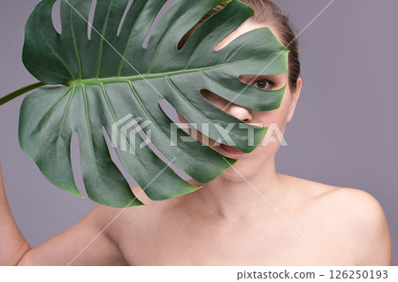 Young caucasian female partially concealed by monstera leaf against neutral 126250193