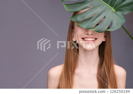 Smiling young caucasian female partially hidden by monstera leaf against purple Smiling young caucasian female partially hidden by monstera leaf against purple 126250194