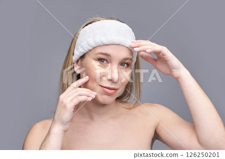 Young caucasian female applying under eye concealer with headband on neutral 126250201