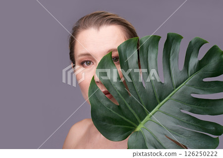 Mature caucasian female partially hidden by large monstera leaf on gray Mature caucasian female partially hidden by large monstera leaf on gray 126250222