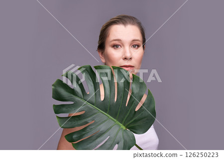 Caucasian adult female posing with monstera leaf against gray background Caucasian adult female posing with monstera leaf against gray background 126250223