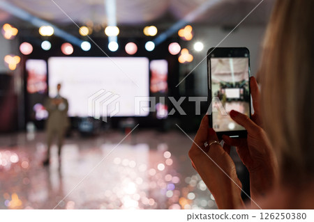 defocus. A woman is filming a man's performance on stage on her smartphone.  126250380