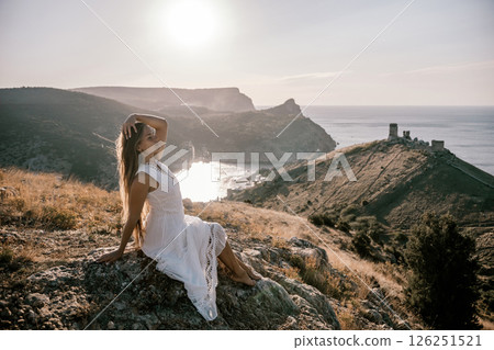 A woman in a white dress is sitting on a rock overlooking a body of water. She is enjoying the view and taking in the scenery. 126251521