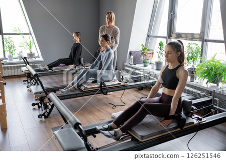 Group of caucasian women exercising with trainer on pilates reformers in a bright studio. Their workout focuses on core strength and flexibility in a modern fitness environment 126251546