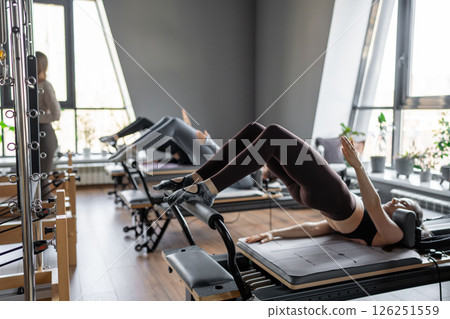A group of woman exercises Glute bridge on a reformer bed machine in natural sunlight, focusing on pelvis and hips. 126251559