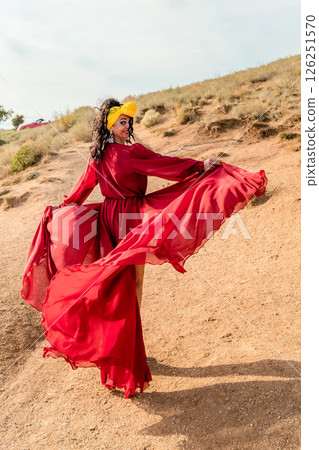 A woman in a red dress is posing for a photo on a dirt road. She is wearing a yellow headband and has her arms outstretched. Concept of freedom and joy. 126251570