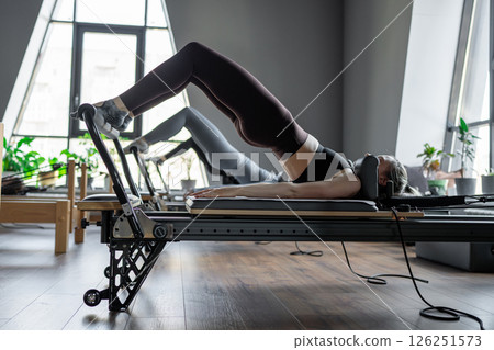 A group of woman exercises Glute bridge on a reformer bed machine in natural sunlight, focusing on pelvis and hips. 126251573