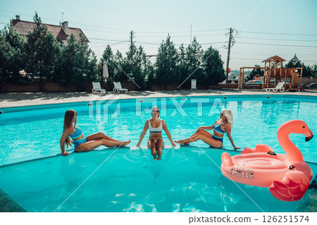 Three women are sitting in a pool, one of them is wearing a pink floatie. Scene is lighthearted and fun, as the women are enjoying their time together in the pool. 126251574