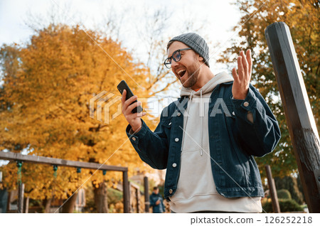 Pleasantly surprised. With smartphone. Beautiful man in warm clothes is outdoors in the park Pleasantly surprised. With smartphone. Beautiful man in warm clothes is outdoors in the park 126252218