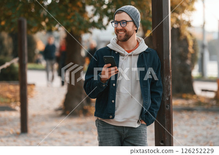 Standing with smartphone. Beautiful man in warm clothes is outdoors in the park Standing with smartphone. Beautiful man in warm clothes is outdoors in the park 126252229