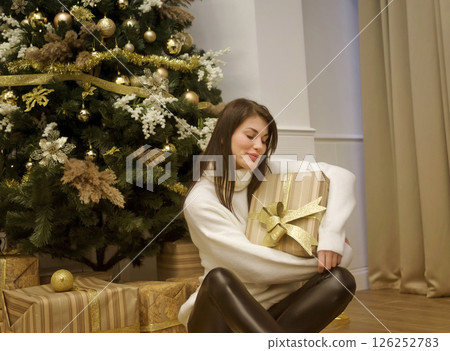 A cute young woman is sitting in front of a Christmas tree and golden boxes with gifts 126252783