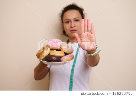 Woman Showing Rejection Gesture Over Plate of Doughnuts Emphasizing Healthy Choices Woman Showing Rejection Gesture Over Plate of Doughnuts Emphasizing Healthy Choices 126253793