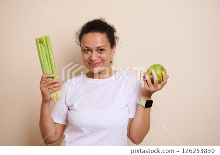Smiling Woman Holding Fresh Green Apple and Celery Stalks Promoting Health and Wellness Smiling Woman Holding Fresh Green Apple and Celery Stalks Promoting Health and Wellness 126253830
