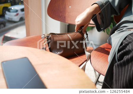 A person adjusts belongings in a brown bag at a sunlit cafe table 126255484