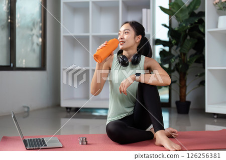 Content Asian woman enjoying water from an orange bottle after a workout, highlighting wellness and fitness recovery at home 126256381