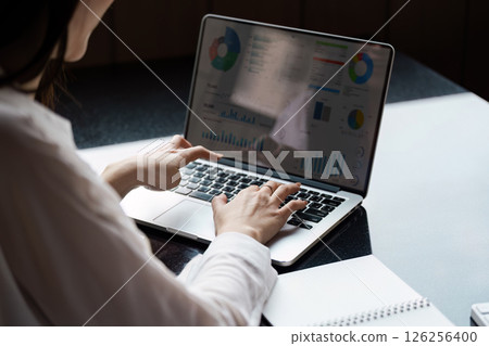 Focused Asian woman analyzing financial data on a laptop while taking notes in a modern office. 126256400