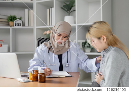 Empathetic female doctor in hijab taking notes while examining a patient in her office. 126256403