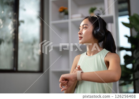 Young woman monitoring fitness progress with smartwatch and headphones during workout at home 126256408