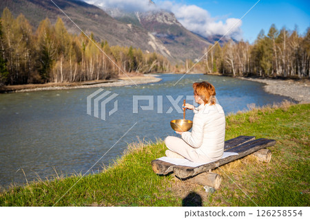 Woman playing a Tibetan singing bowl by a mountain river in Altai. Concept of sound healing, spiritual alignment, and deep connection with natural elements 126258554