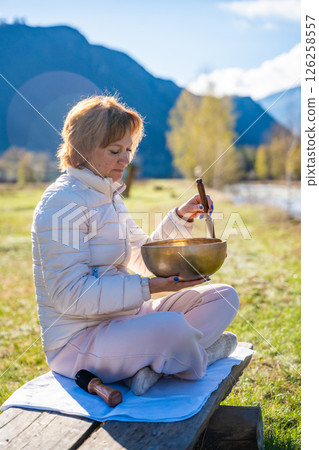 Woman playing a Tibetan singing bowl by a mountain river in Altai. Concept of sound healing, spiritual alignment, and deep connection with natural elements 126258557