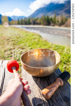 Point of view of hand playing a Tibetan singing bowl near mountain river in Altai. Concept of personal ritual, sound healing, and spiritual harmony with wild nature. 126258563