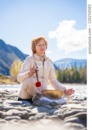 Woman performing a sound ritual with a Tibetan singing bowl by the Altai river. Concept of feminine energy, sacred vibration, and shamanic connection to the Earth Woman performing a sound ritual with a Tibetan singing bowl by the Altai river. Concept of feminine energy, sacred vibration, and shamanic connection to the Earth 126258565