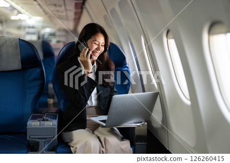Business travel with a young professional woman working on a laptop and talking on the phone in an airplane Business travel with a young professional woman working on a laptop and talking on the phone in an airplane 126260415