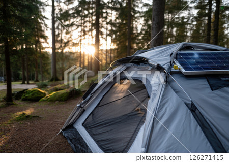 Tent equipped with solar panel is set up in serene forest, capturing warm glow of sunset filtering through trees. peaceful setting highlights sustainable camping practices 126271415