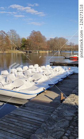 Trakai Lithuania - 03.28.2025 White pedal boats docked on a wooden pier beside a calm lake with a wooden bridge and autumn trees under a clear blue sky Trakai Lithuania - 03.28.2025 White pedal boats docked on a wooden pier beside a calm lake with a wooden bridge and autumn trees under a clear blue sky 126273118